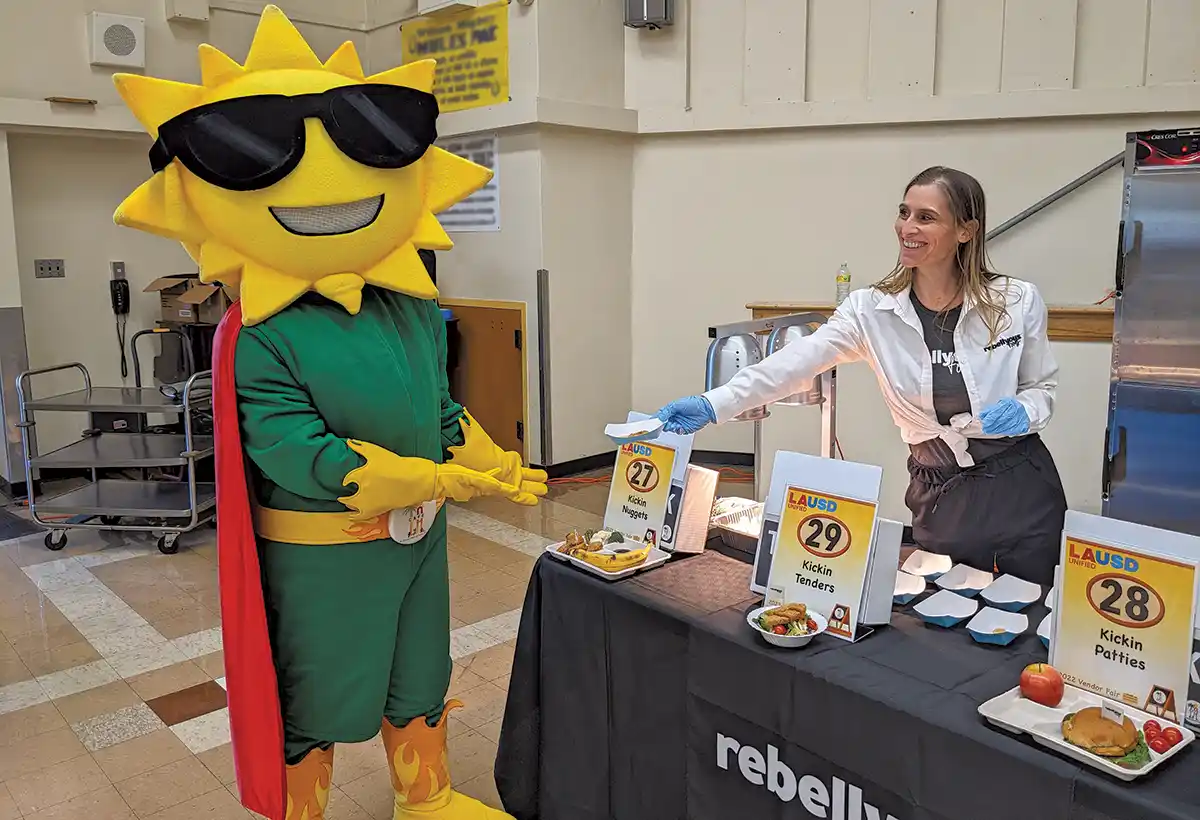 Kristie Middleton extends a food sample to “Ray,” the mascot for Los Angeles Unified School District’s food services department, “Cafe LA” at a student taste testing where students were able to sample the Rebellyous Foods products. 
